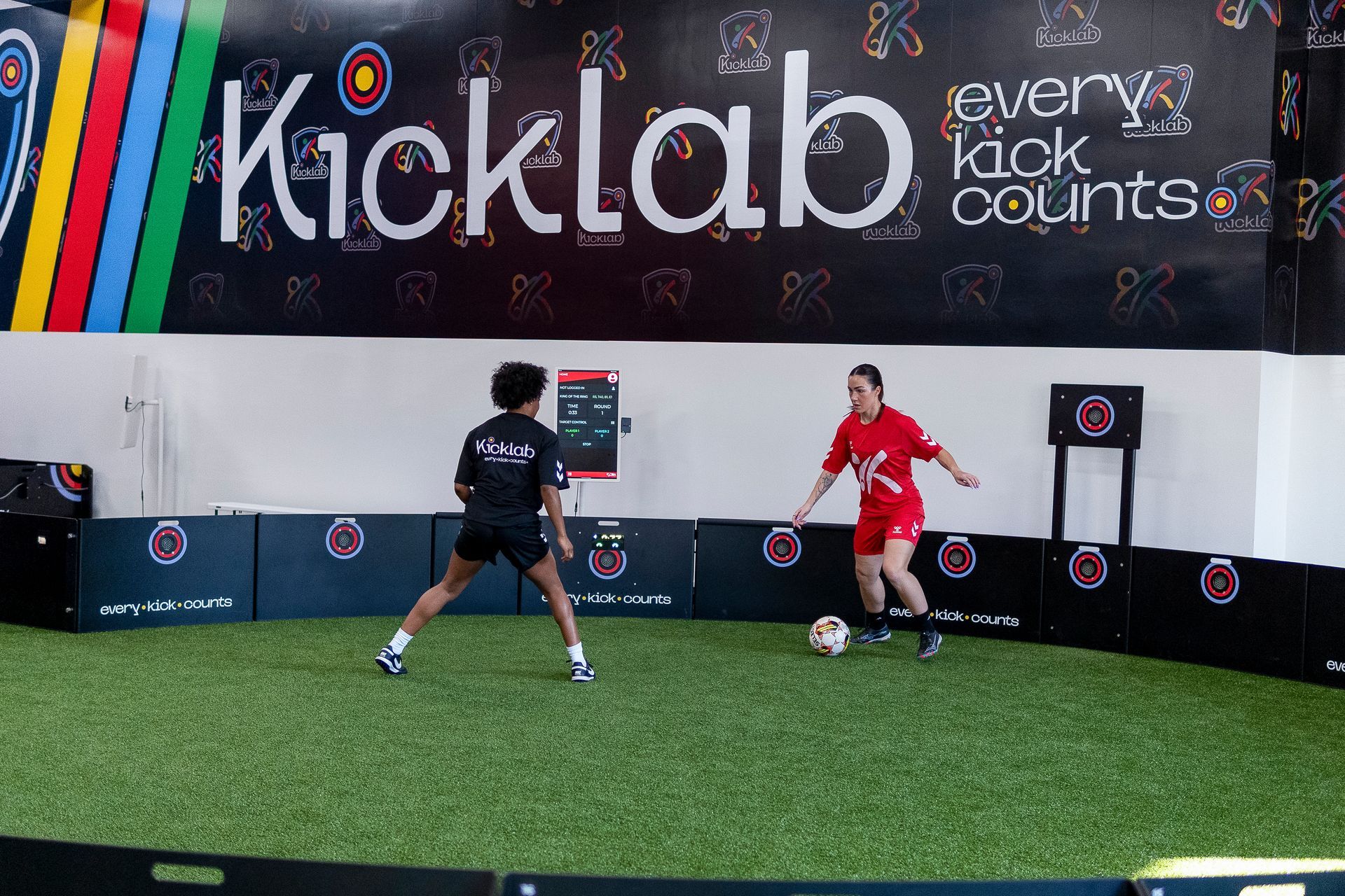 Two women are playing soccer in front of a sign that says kicklab