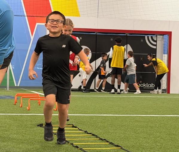 A child in a black t-shirt and shorts performs agility ladder exercises on an indoor turf field with others in the back.