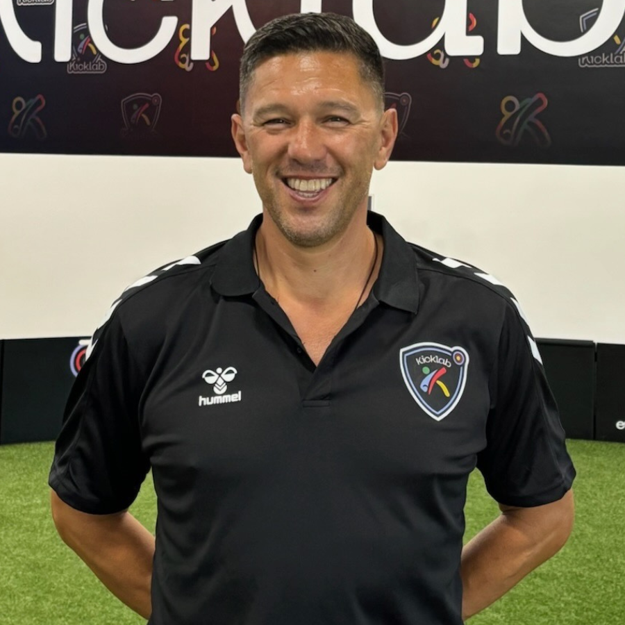 Man in black polo shirt with logo, smiling, arms crossed. Stands in a KickLab studio.