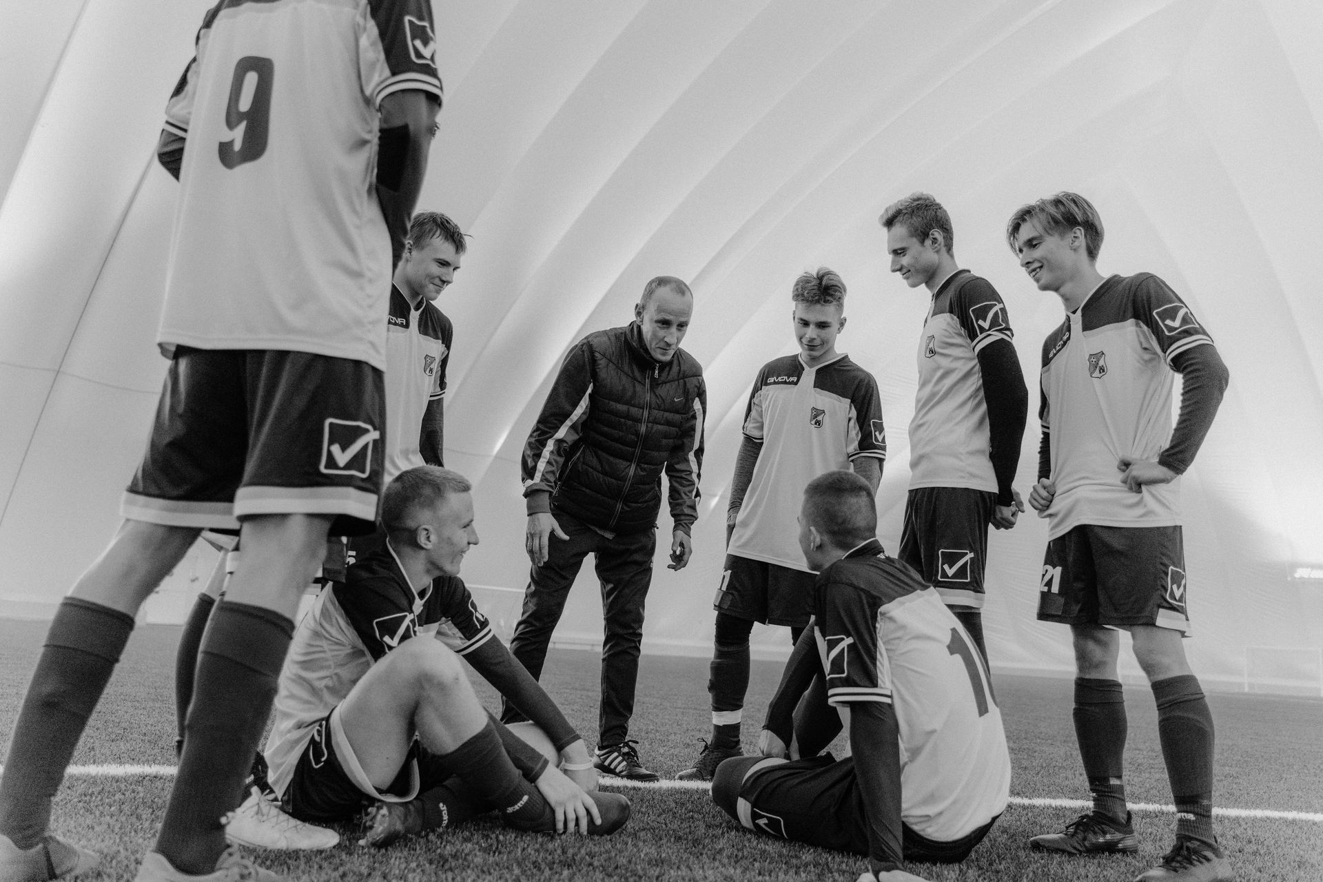 Soccer team huddle in an indoor white dome. Coach speaks to players, some standing, others sitting.