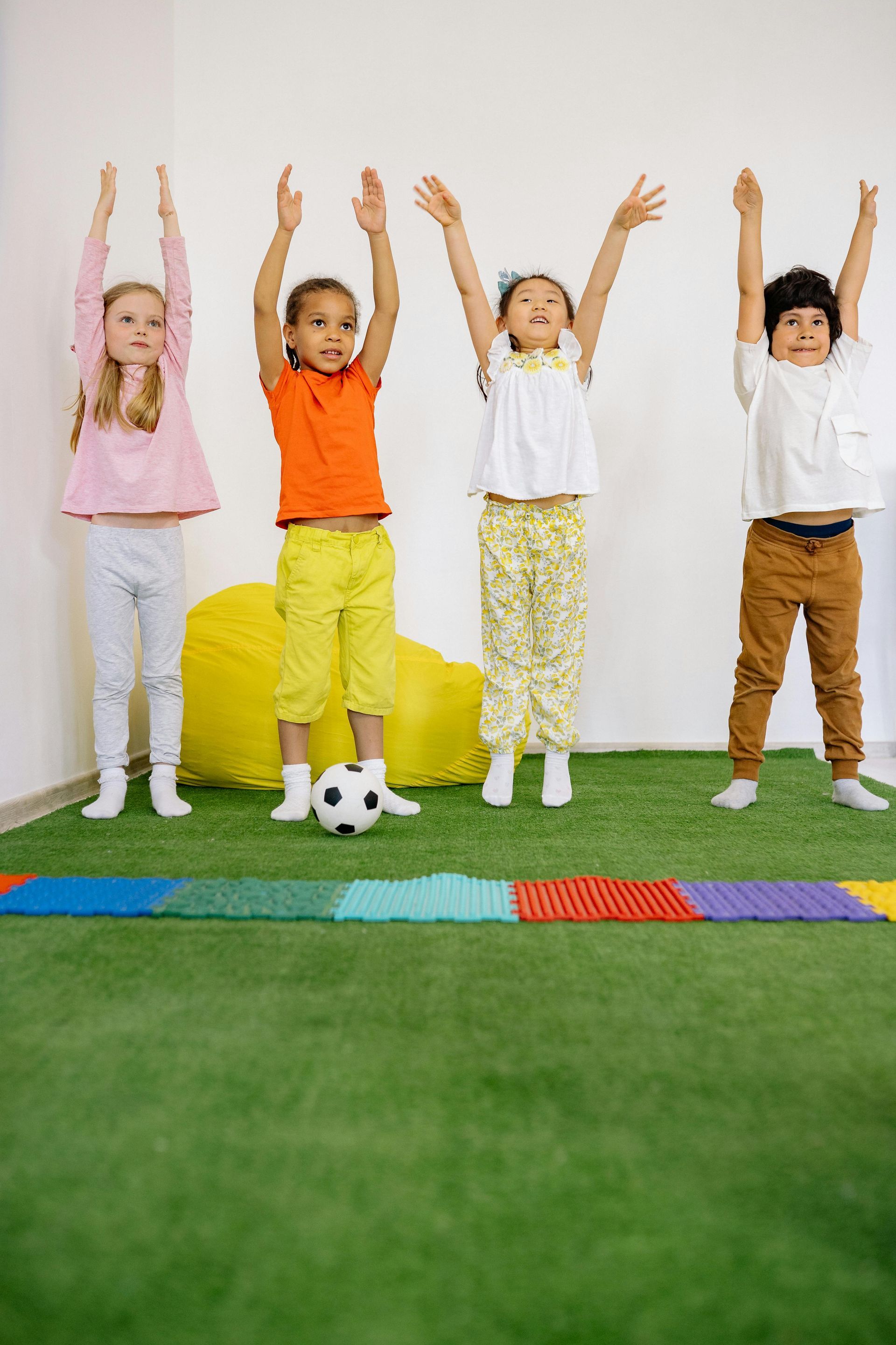 Four children with arms raised, standing on green turf near a soccer ball and colorful mat.