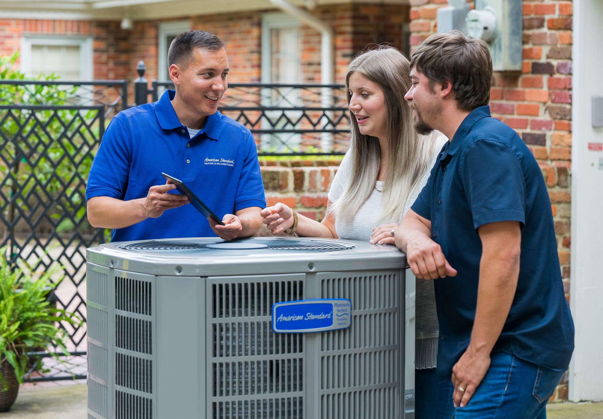 HVAC technician showing a tablet to a couple next to an air conditioning unit outside a brick house.