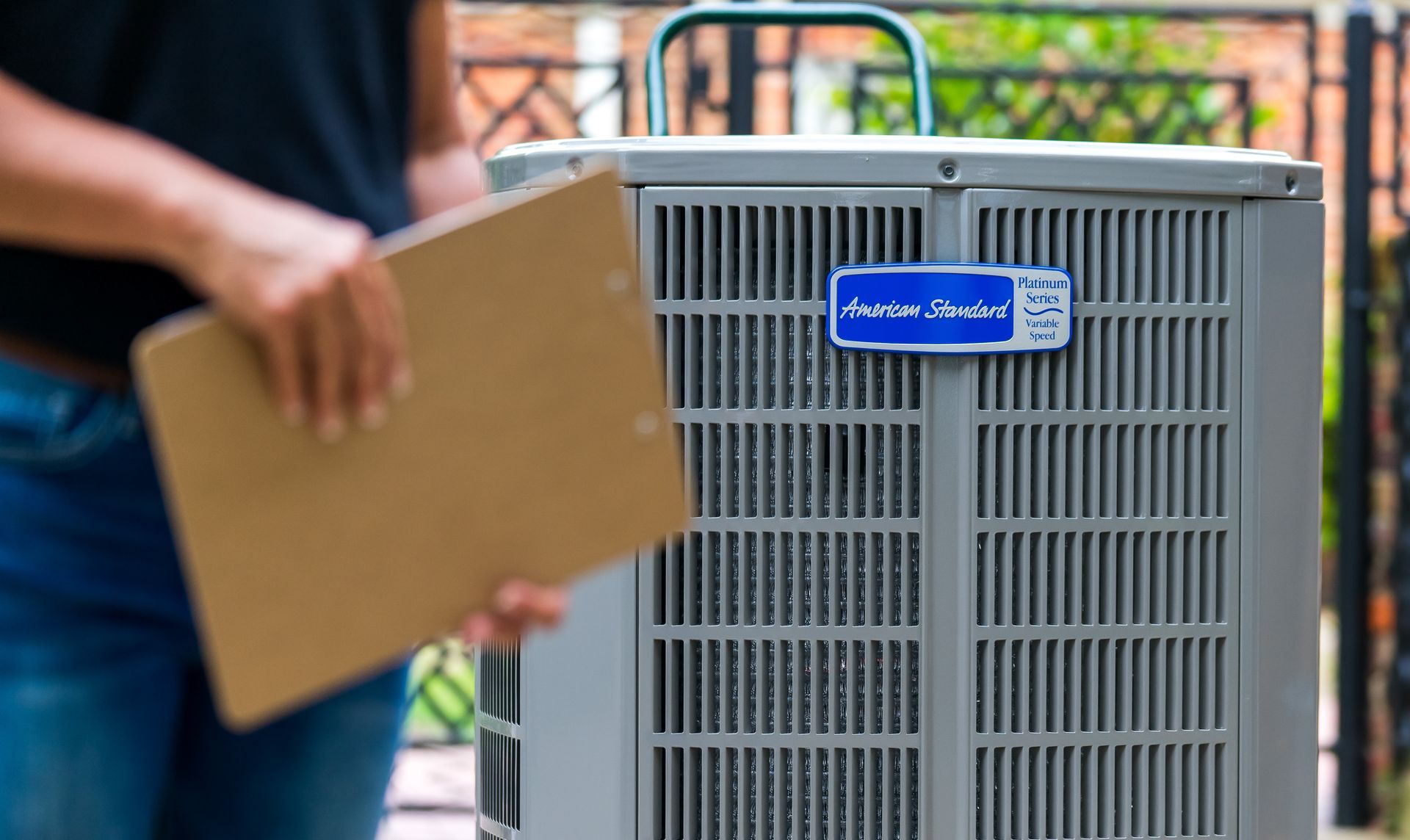 Person holding clipboard next to an American Standard air conditioning unit outdoors.