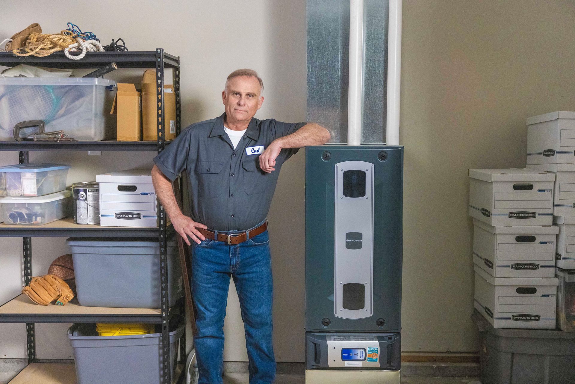 Man in work clothes poses next to a dark gray heating unit in a garage, leaning on it.
