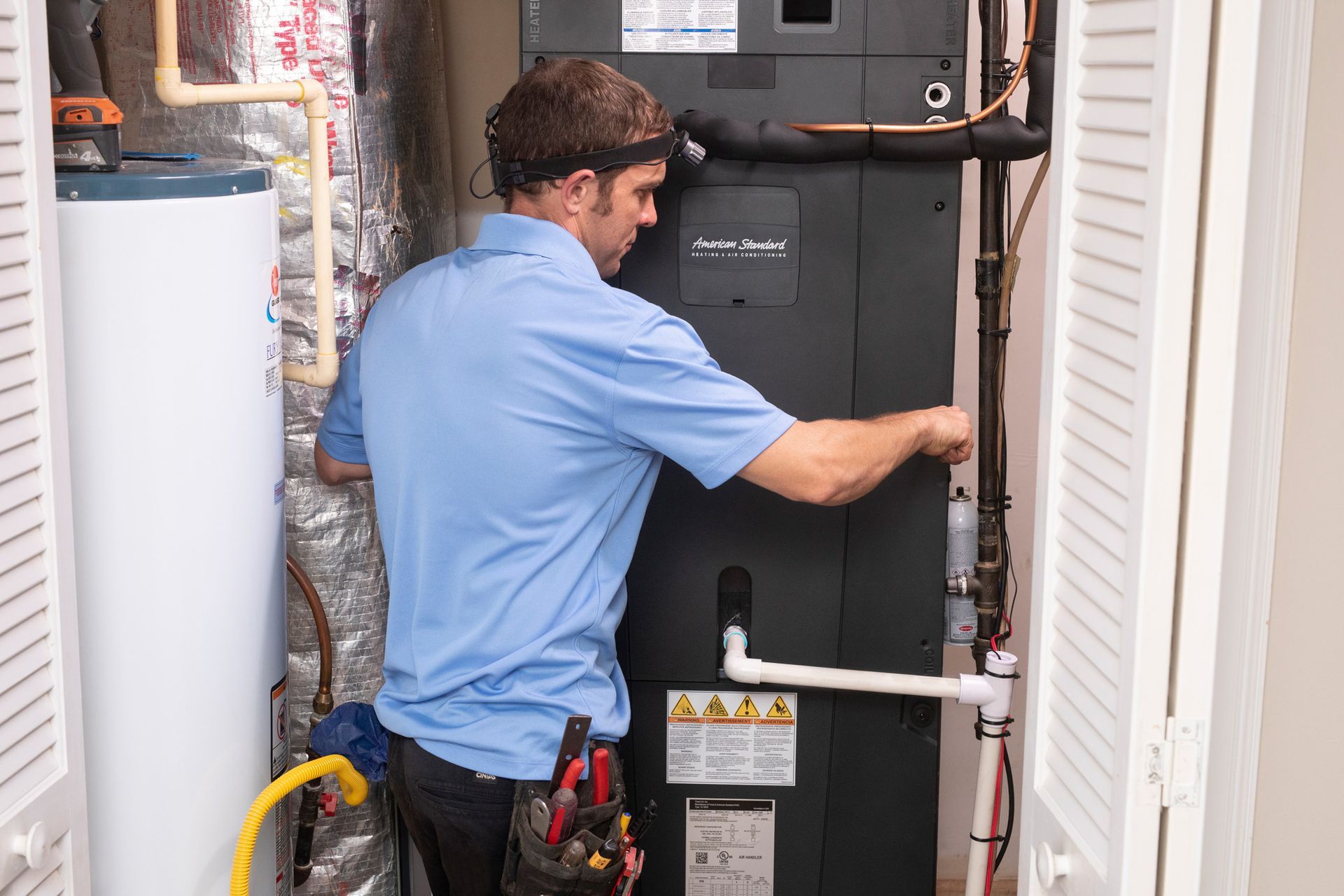 HVAC technician inspecting an air handler unit inside a utility closet; water heater visible.