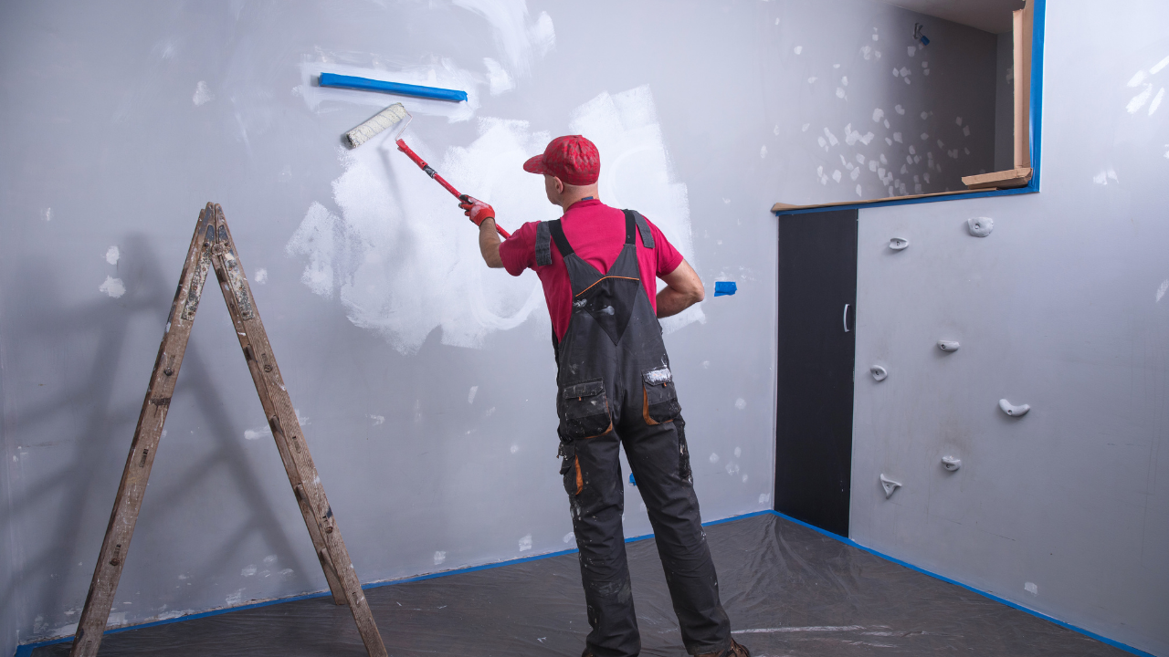 A person in a red shirt and dark overalls uses a paint roller to apply white paint to a grey wall in a room.