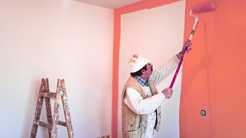 A person wearing a hat and vest uses a long-handled roller to paint a wall bright orange in a room with a wooden ladder.