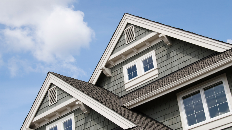 Gable roofs of a grey shingled house against a bright blue sky with clouds.