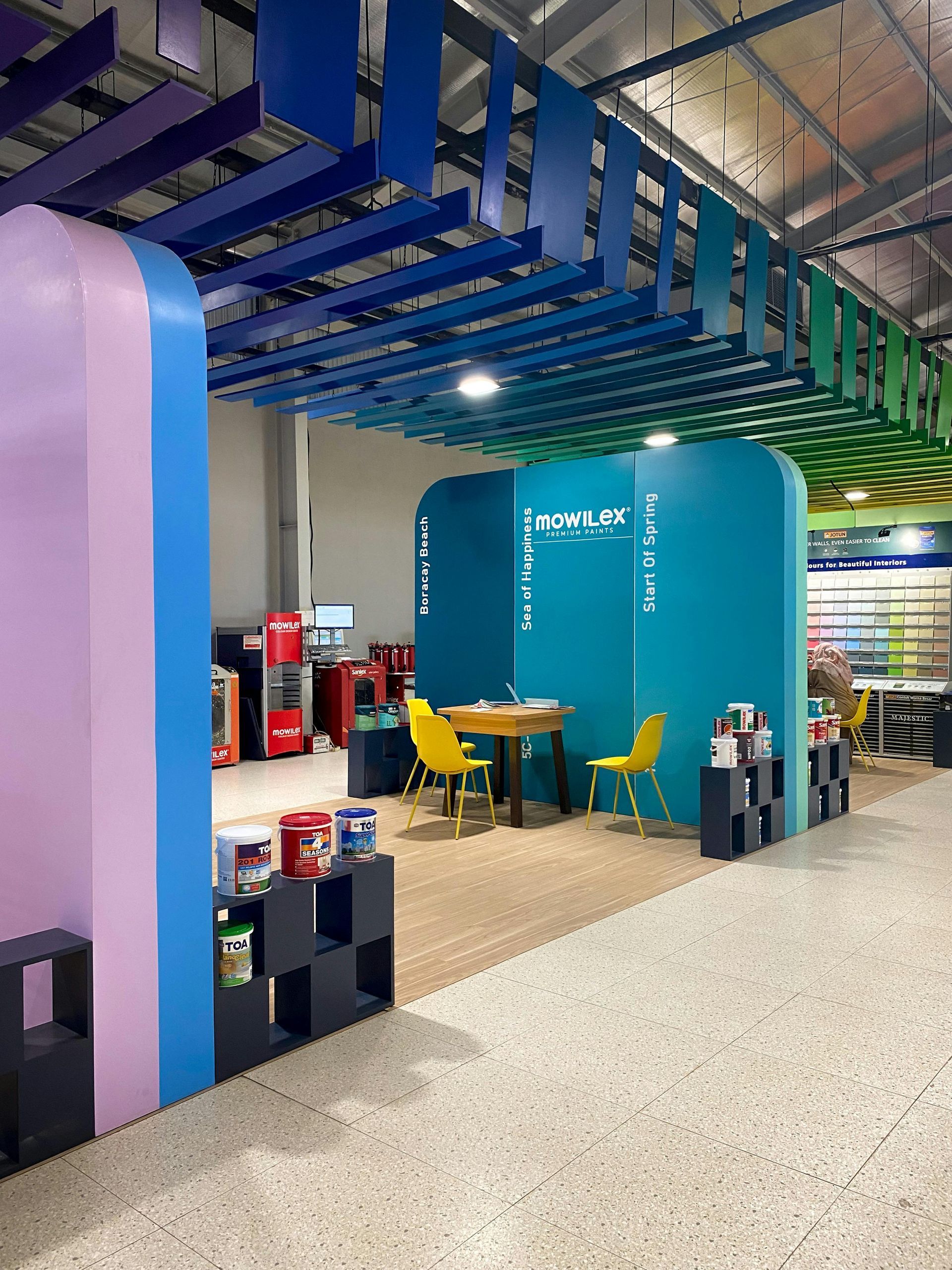 A colorful, modern retail display with blue and pink walls, a central seating area with yellow chairs, and ceiling baffles.