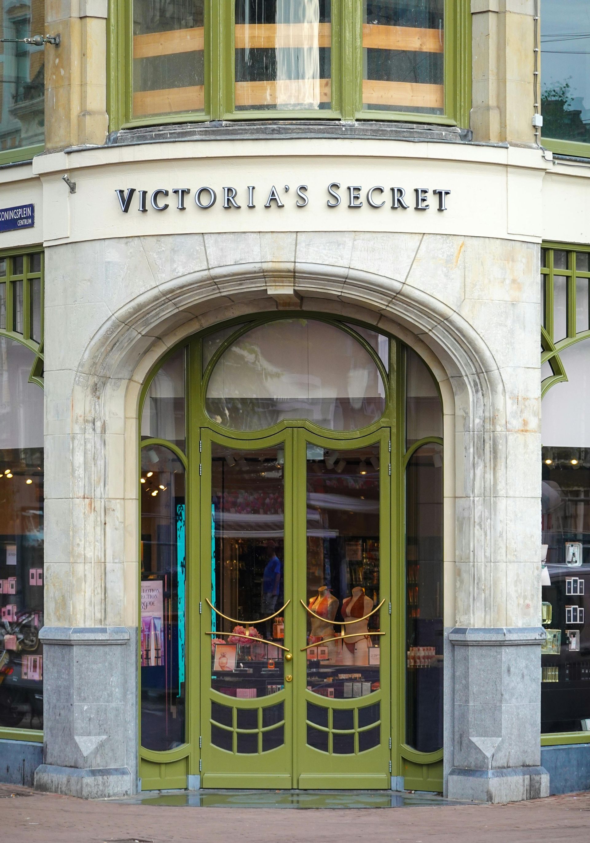 The exterior entrance of a Victoria's Secret store featuring an ornate, arched, light green doorway and stone architecture.