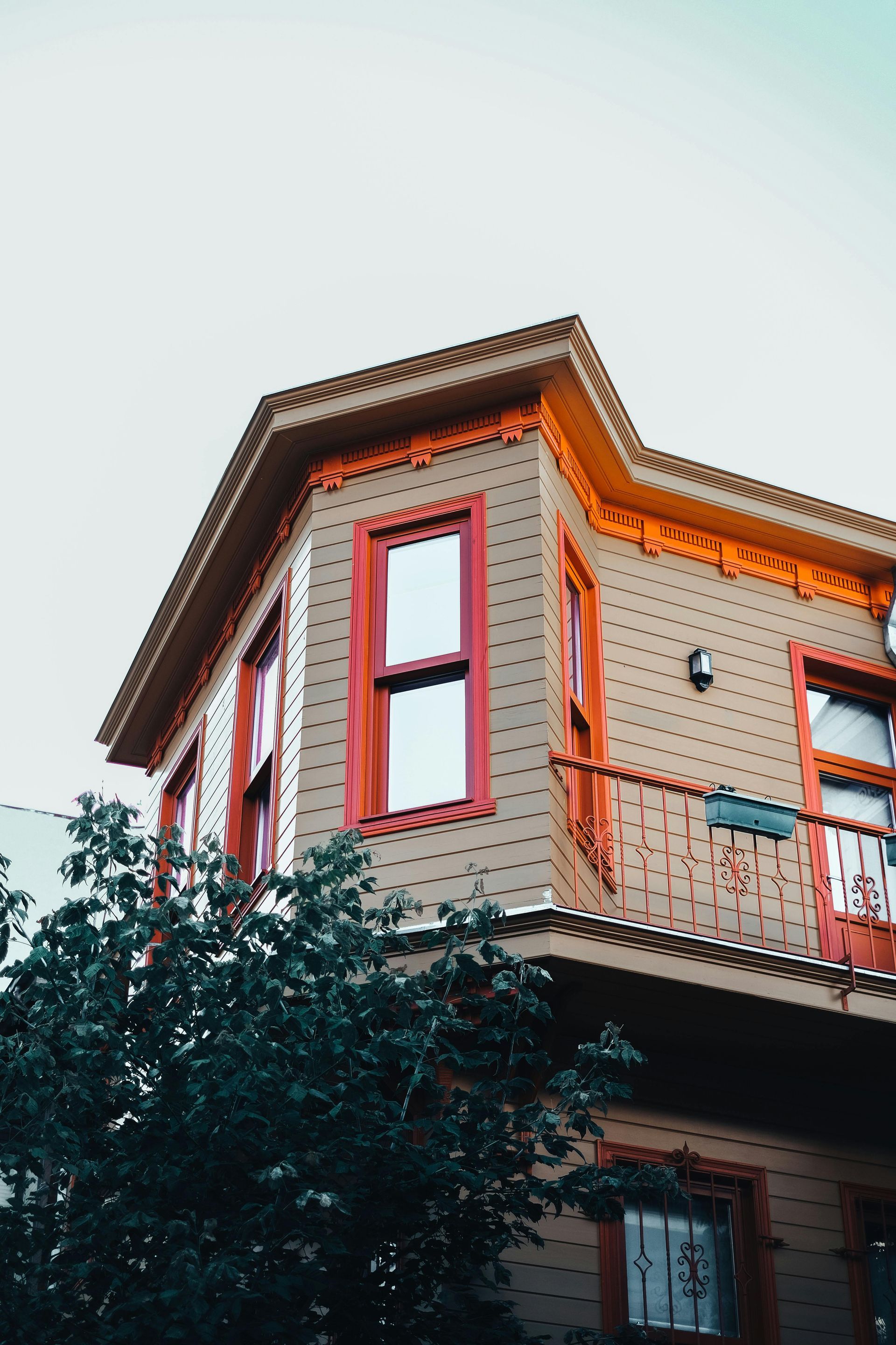 A beige Victorian-style building features bright orange trim, a bay window, and a small balcony, framed by green foliage.
