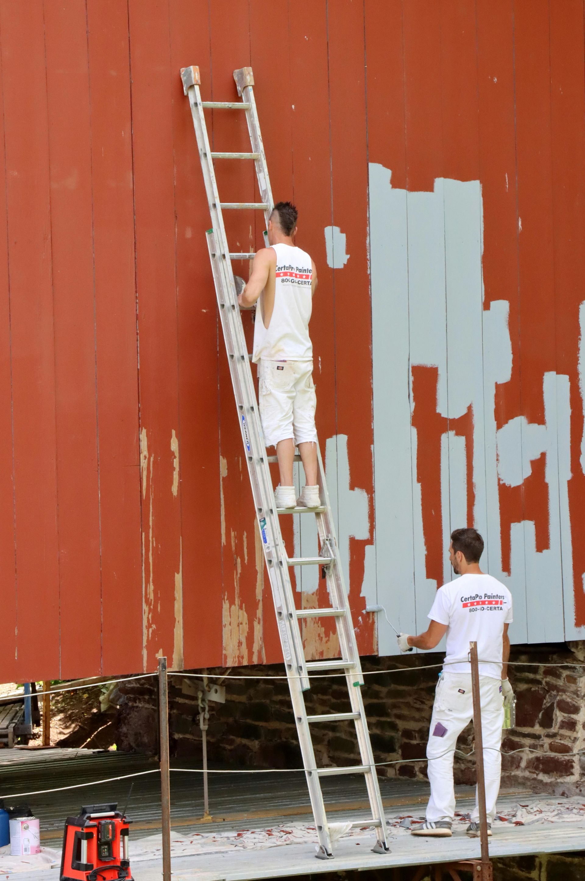 Two people in white work clothes paint a red wall with light blue paint, one standing on a ladder and one on the ground.