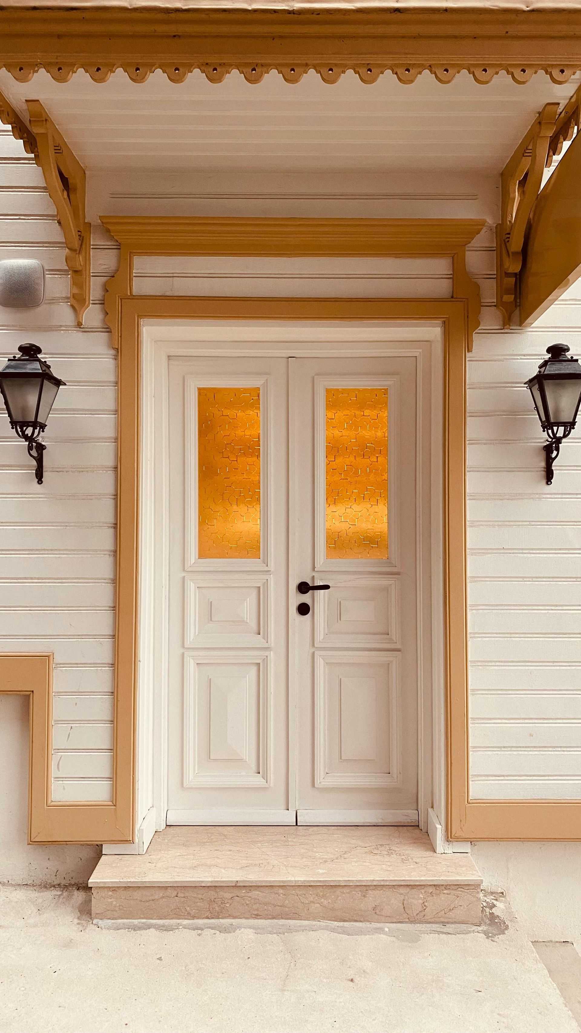 White double doors with golden glass panels and ornate trim under a canopy, flanked by two outdoor wall lanterns.
