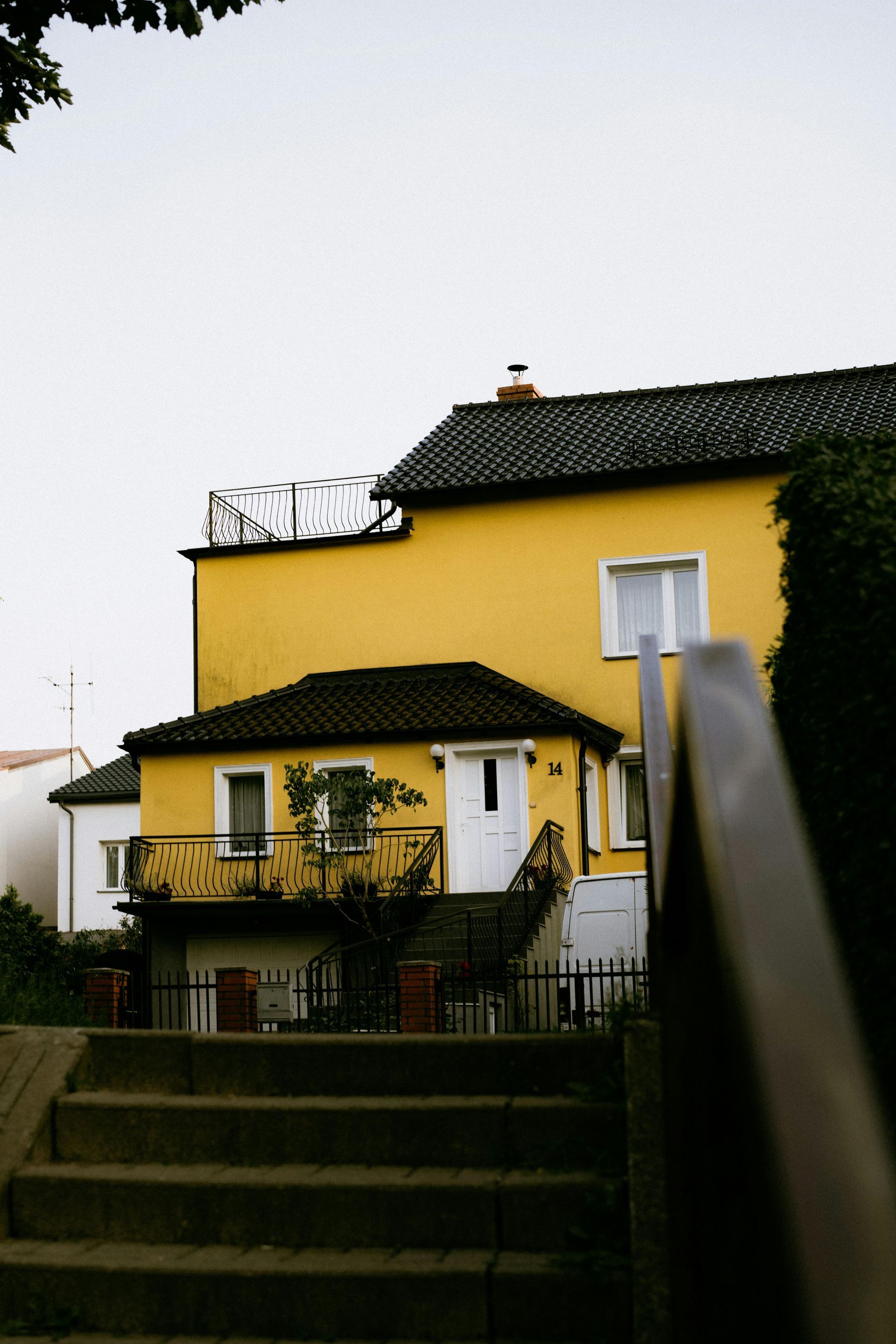 A bright yellow two-story house with a dark tiled roof, seen from the bottom of outdoor concrete stairs.