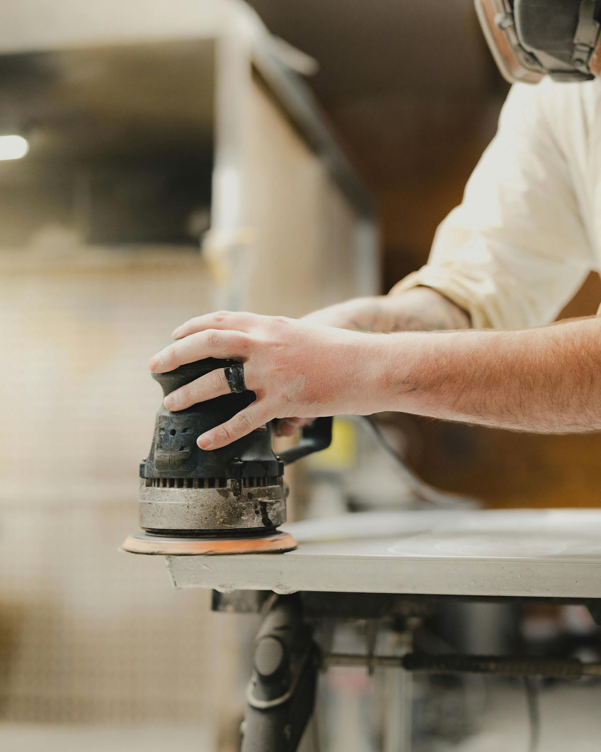 A person wearing safety glasses uses a handheld sander to smooth the surface of a light-colored wooden table in a workshop.