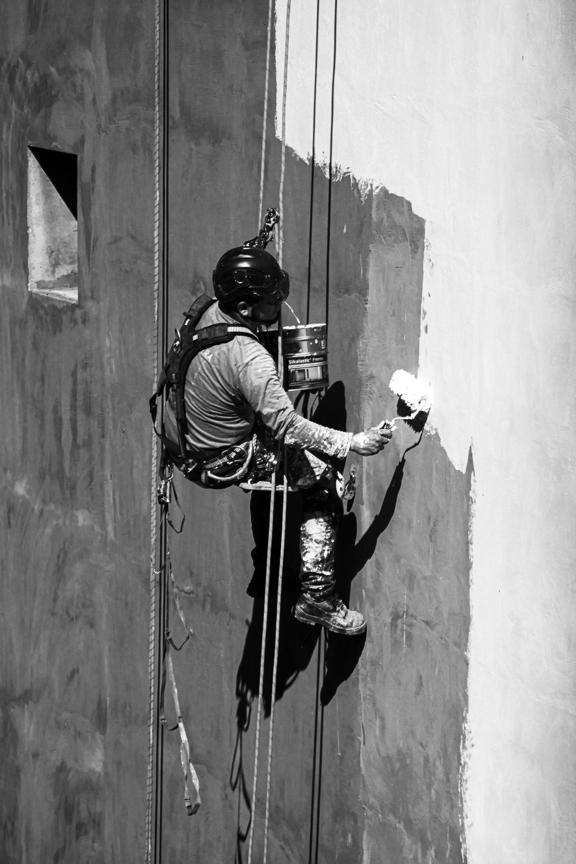 A person suspended by ropes against a tall wall, using a roller to paint a section of the surface white.
