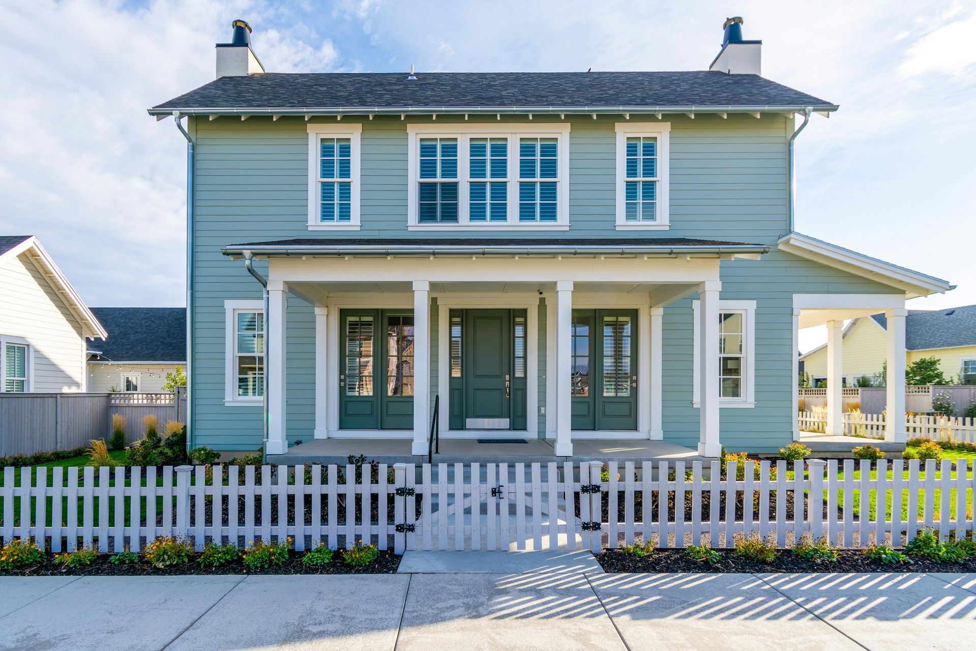 A light blue two-story house with a white picket fence, a covered front porch, and a sidewalk in front.