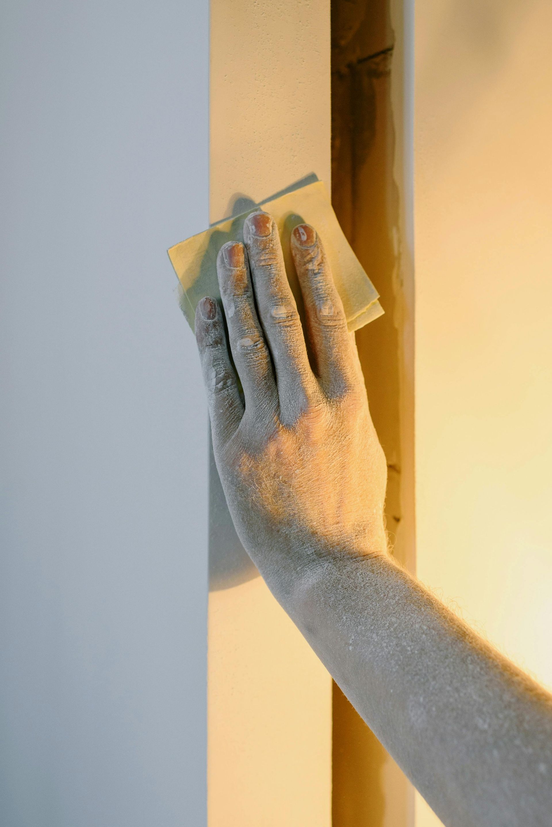 A hand covered in drywall dust uses a sanding block to smooth the surface of a wall corner.