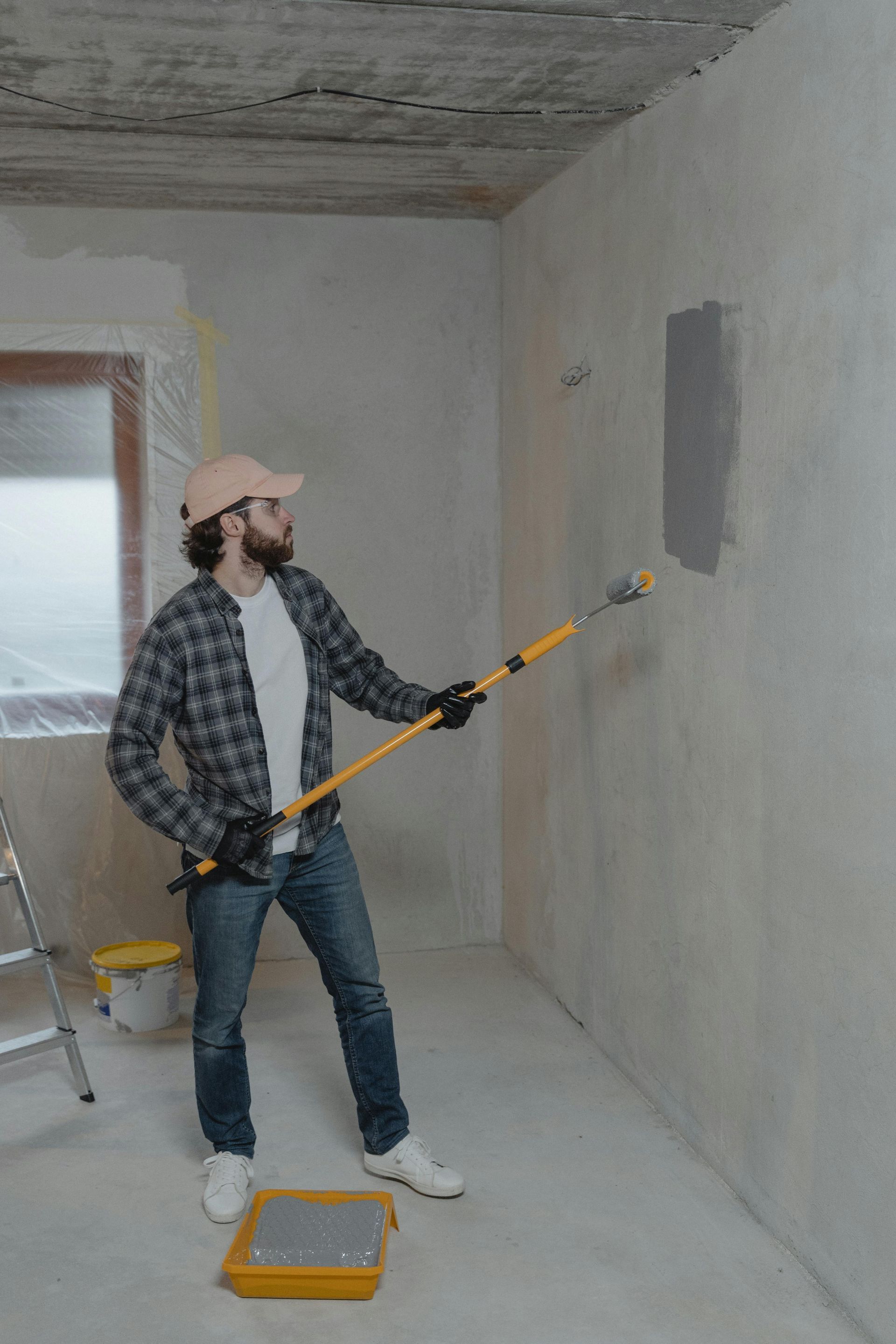 A person in a plaid shirt and cap uses a long-handled roller to paint a small gray patch on a light-colored wall.