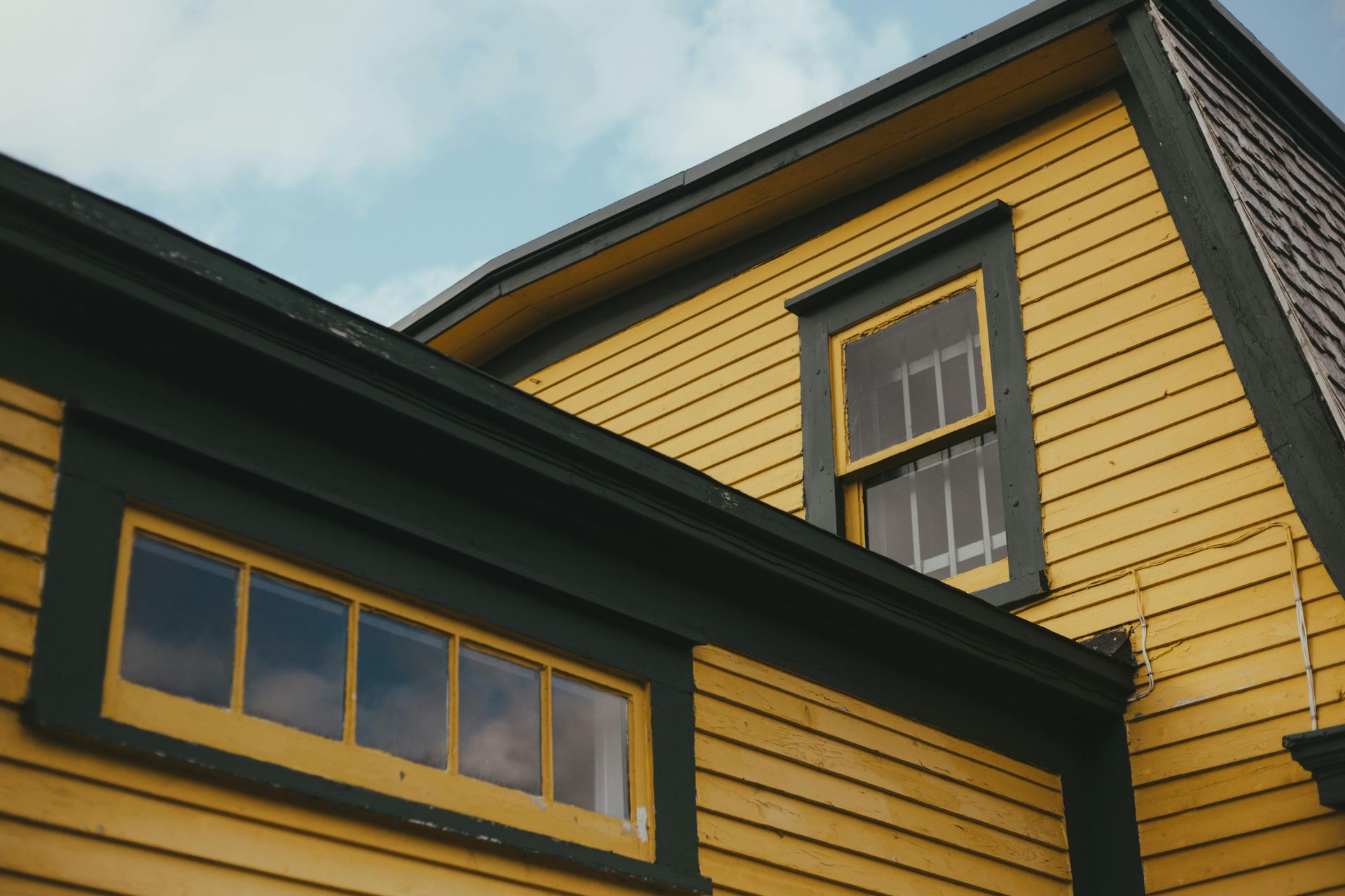 A low-angle view of a yellow house with dark green trim, featuring a horizontal rectangular window and an upper window.
