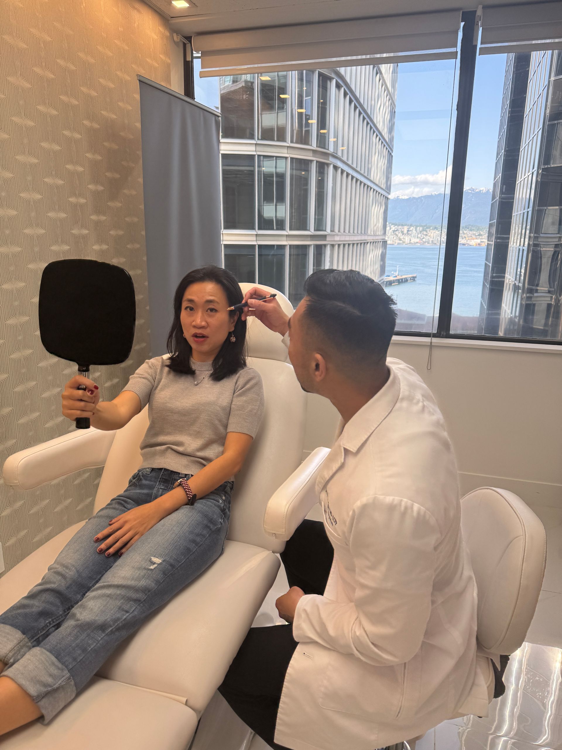 Woman in jeans holds mirror, doctor examines her face, room with city/water view.