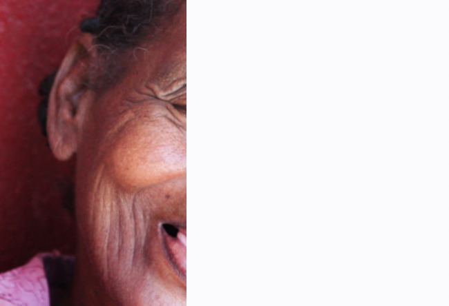 Close-up of an elderly woman with deep wrinkles, smiling with pink shirt, against a red background.