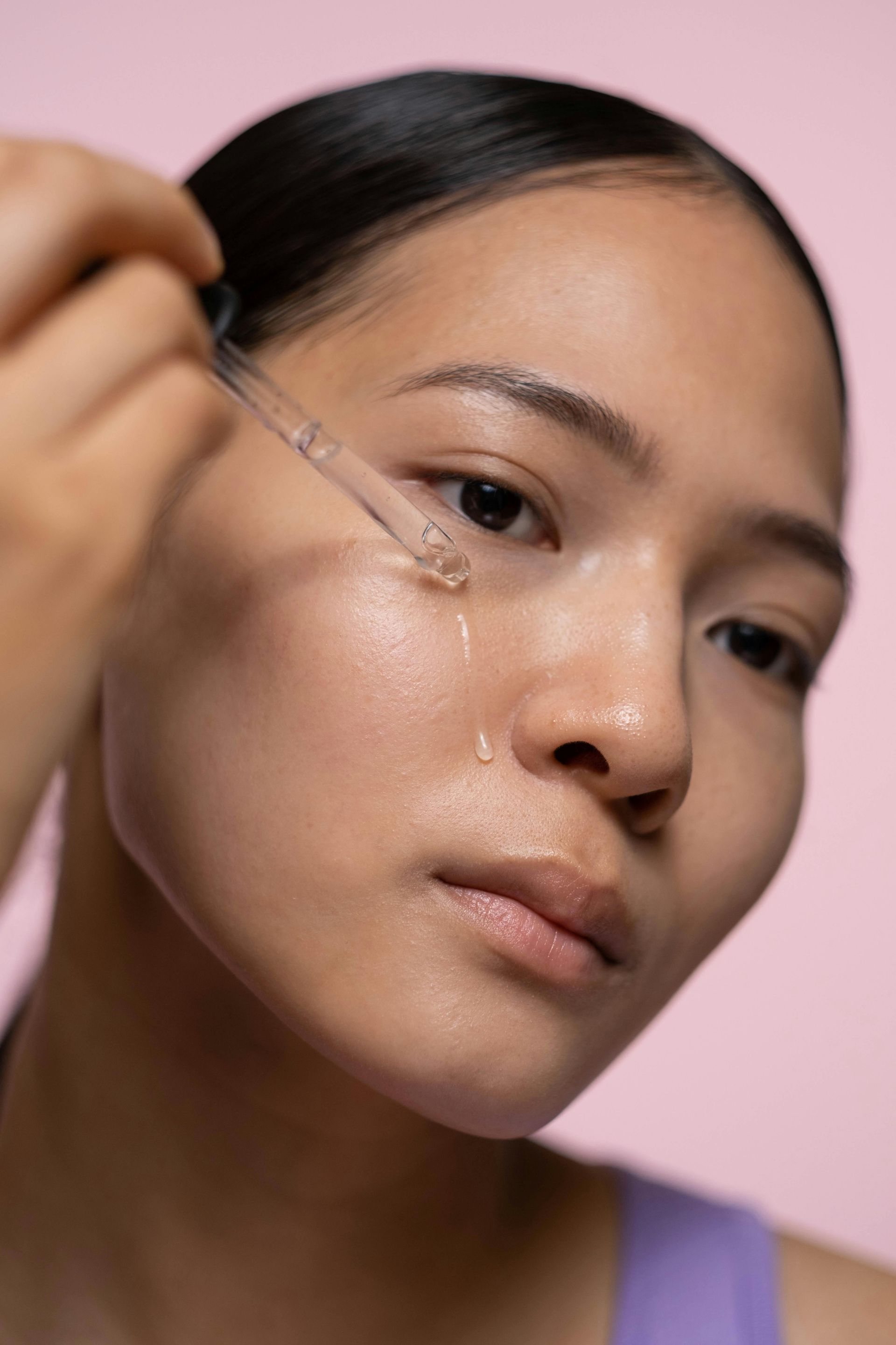 Woman applying skincare serum with dropper on cheek, pink background.