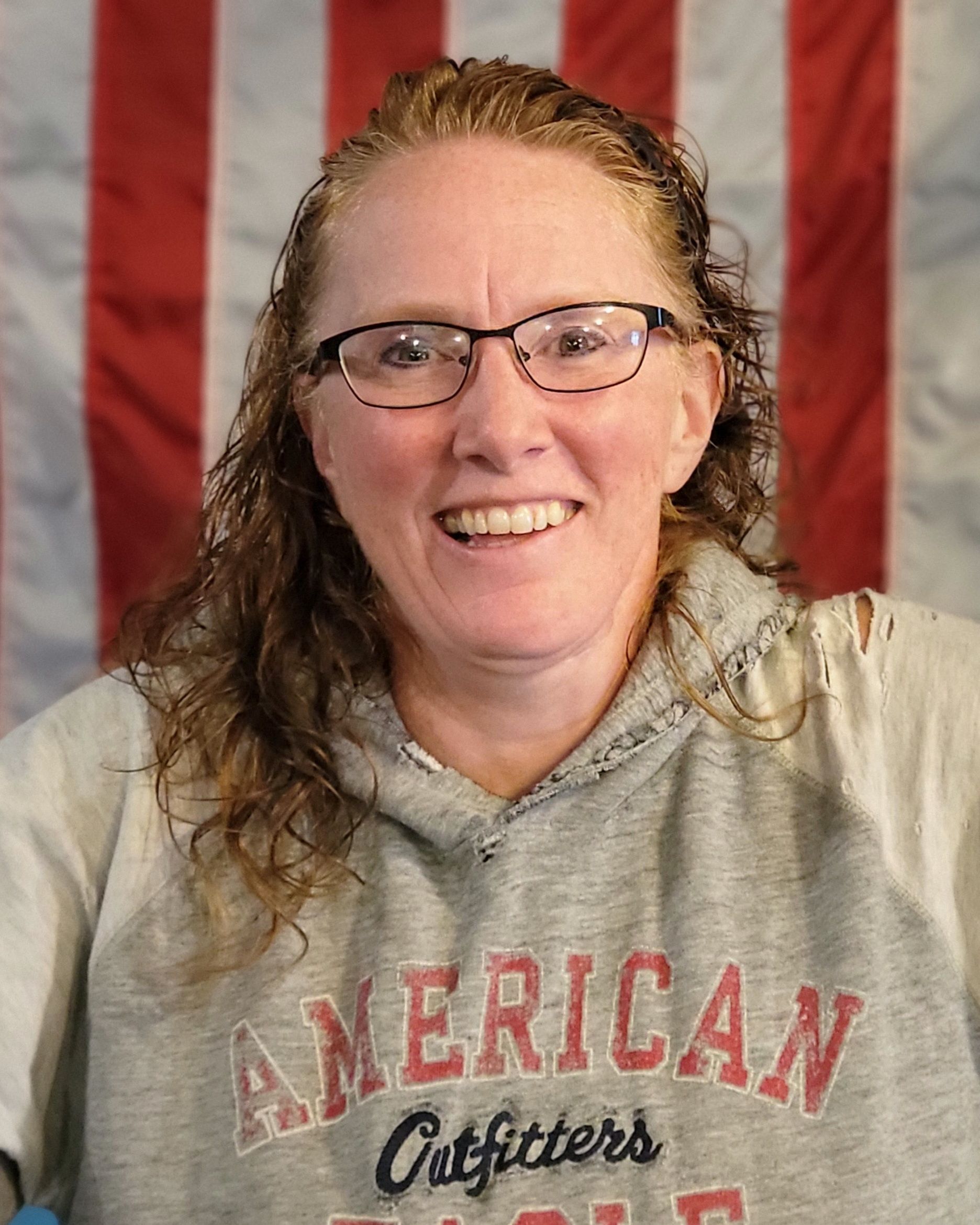 Woman wearing glasses and a gray sweatshirt smiles in front of an American flag.