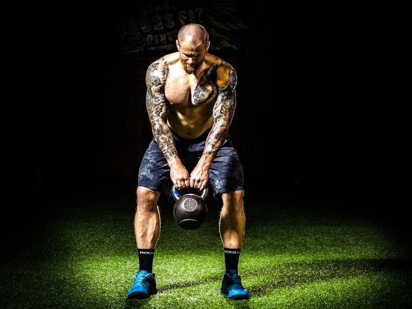 Man with tattoos performs a kettlebell exercise on a green, lit surface.