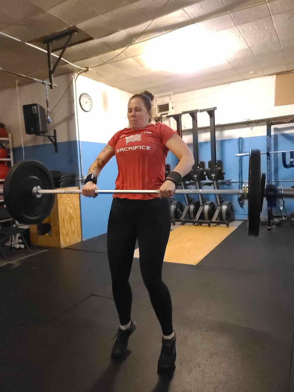 Woman in red shirt performs barbell power clean in a gym. She is airborne, holding the bar at chest level.