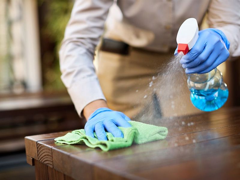 Person wearing blue gloves spraying cleaner on a wooden table with a green cloth.