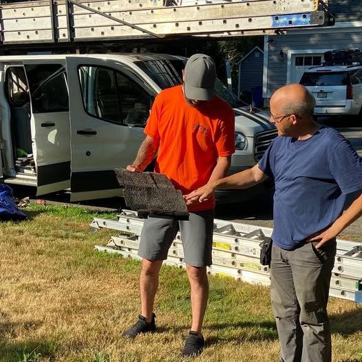 Two men examining a damaged roof shingle, near a work van and ladders on a lawn. One man wears an orange shirt.