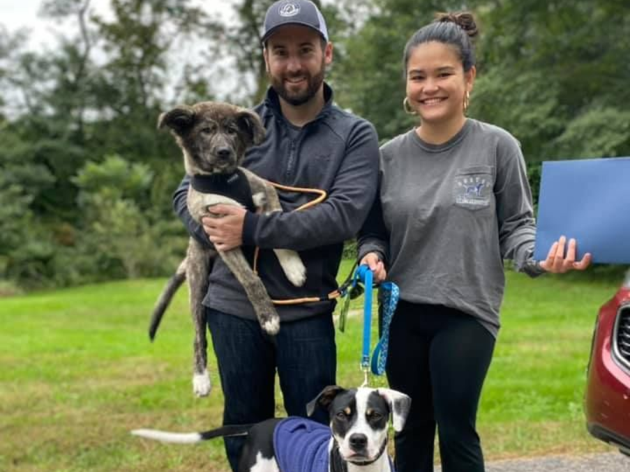 Couple with two dogs outdoors; one held, one on a leash, by a car.
