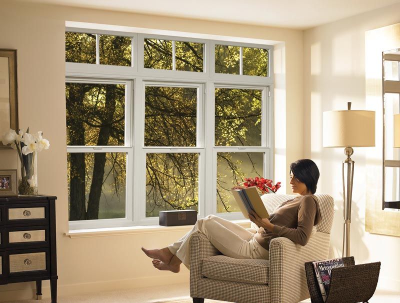 Woman reading by a large window, looking at fall trees outside. Interior setting with armchair, lamp, and cabinet.