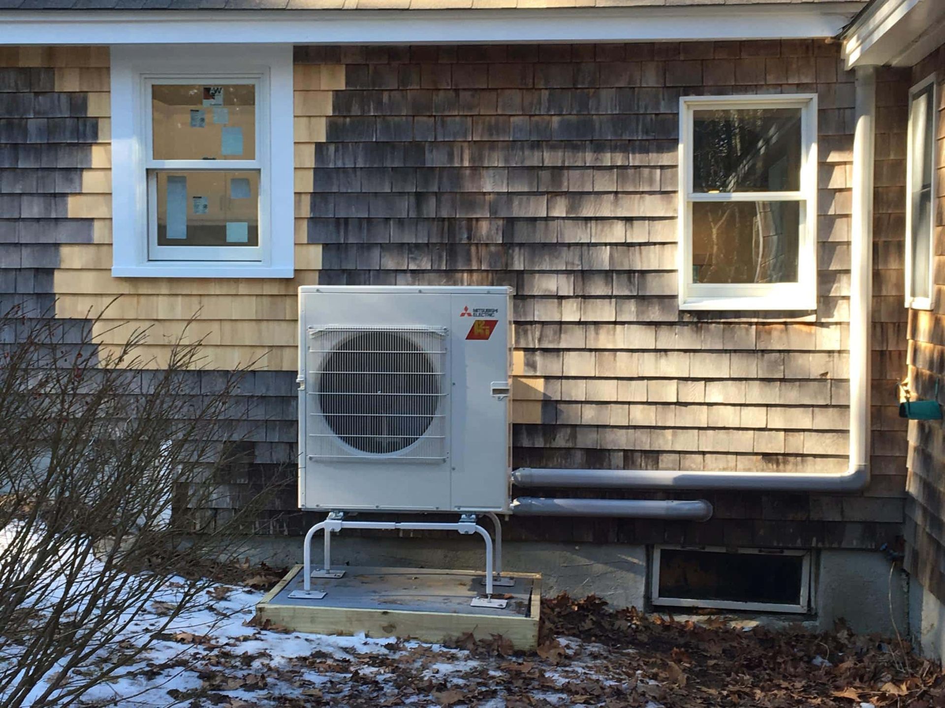 An outdoor heat pump unit on a stand, mounted on a wood-shingled house exterior near windows and a small window well.