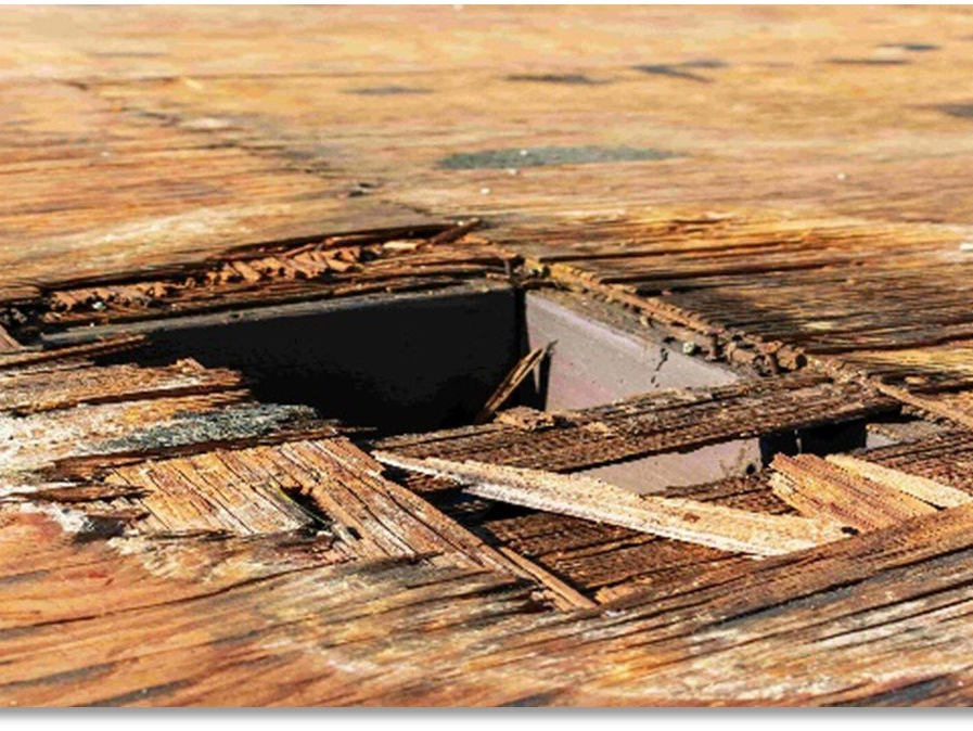 Damaged wooden floor with a square hole exposing dark interior. Boards are splintered and weathered.