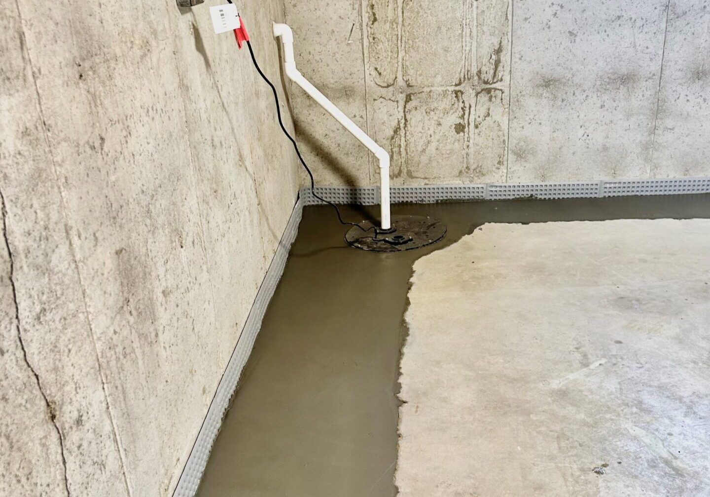 Sump pump in a basement, with a concrete floor and walls. A white pipe extends upwards.