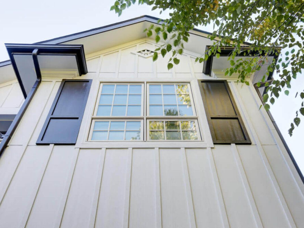 White house exterior with dark shutters, windows, and roof trim. Green tree branches in the upper right.