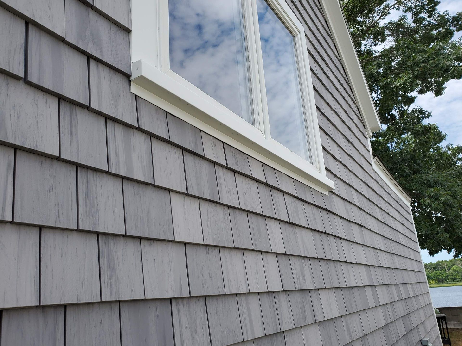 Gray shingle siding on a house, white-framed window reflecting clouds.