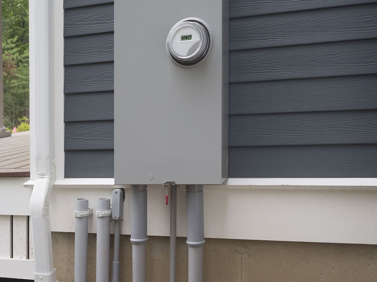 Gray electrical meter box on a house with gray siding and white trim. Gray conduit pipes below.