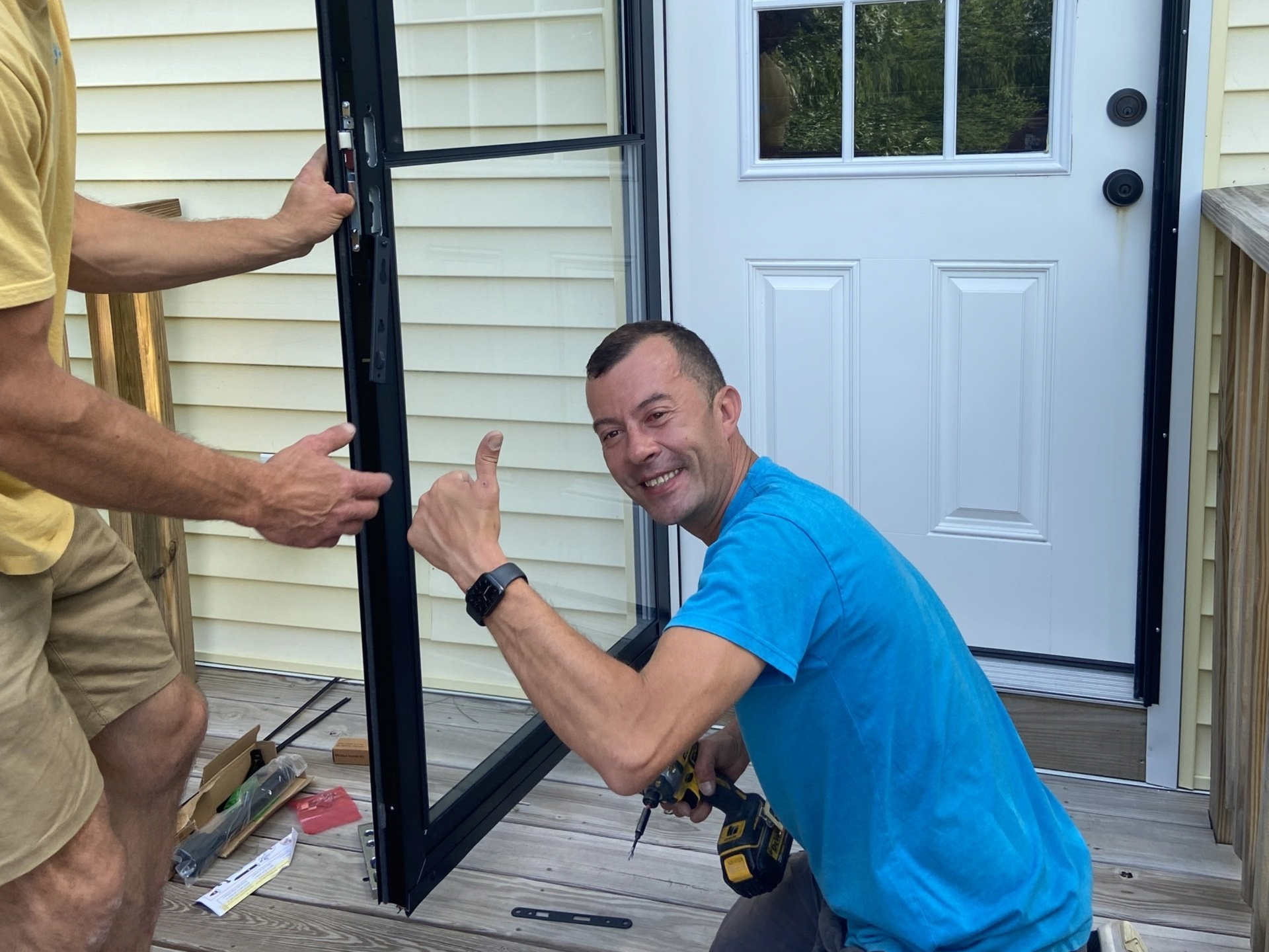Two people installing a black screen door on a porch; one gives a thumbs up while holding a drill.