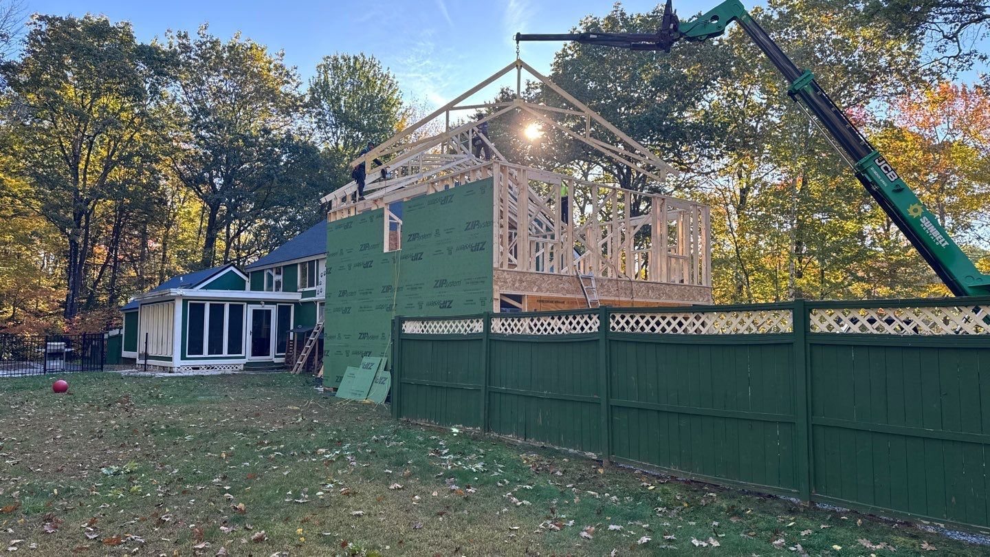 House addition under construction; crane lifting roof frame. Green siding, fence, and equipment. Autumn trees in background.