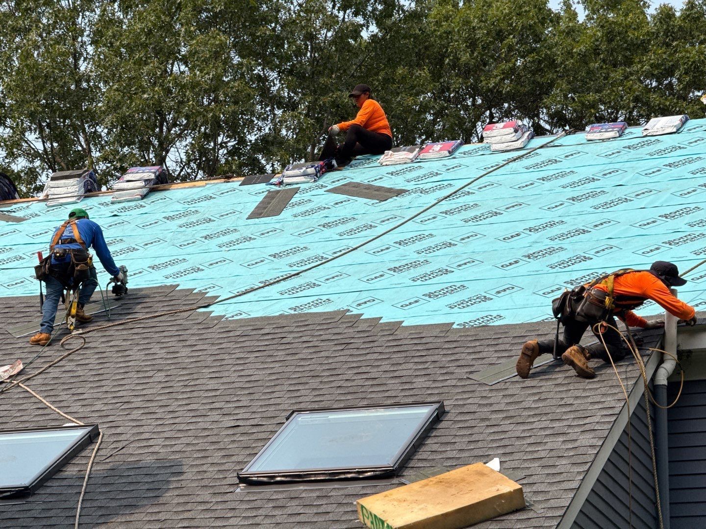 Roofers installing shingles on a house; sky lights visible, three workers are on the roof.