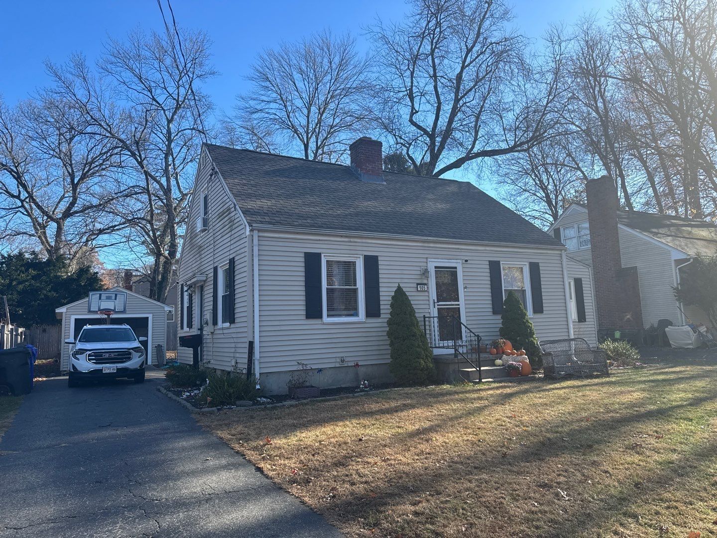 Small beige house with dark shutters, driveway, and detached garage on a sunny day.