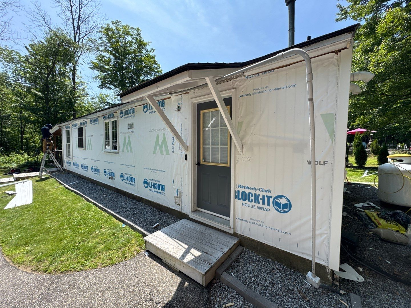 Exterior of a small building under construction, wrapped in blue sheathing with a wooden door and small windows.