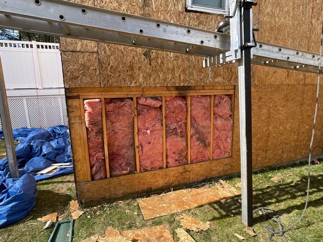 Exterior wall under construction with exposed insulation, wood framing, and ladder.