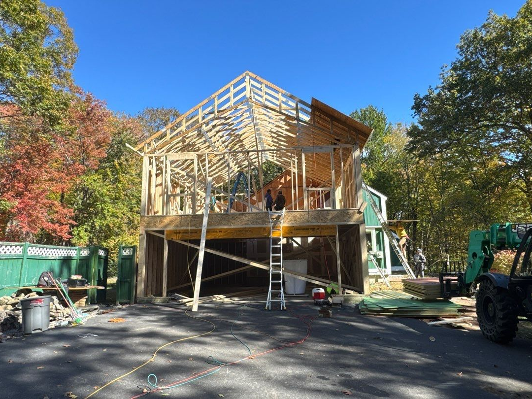 Construction site: wooden frame of a house in progress; trees with fall foliage in background.