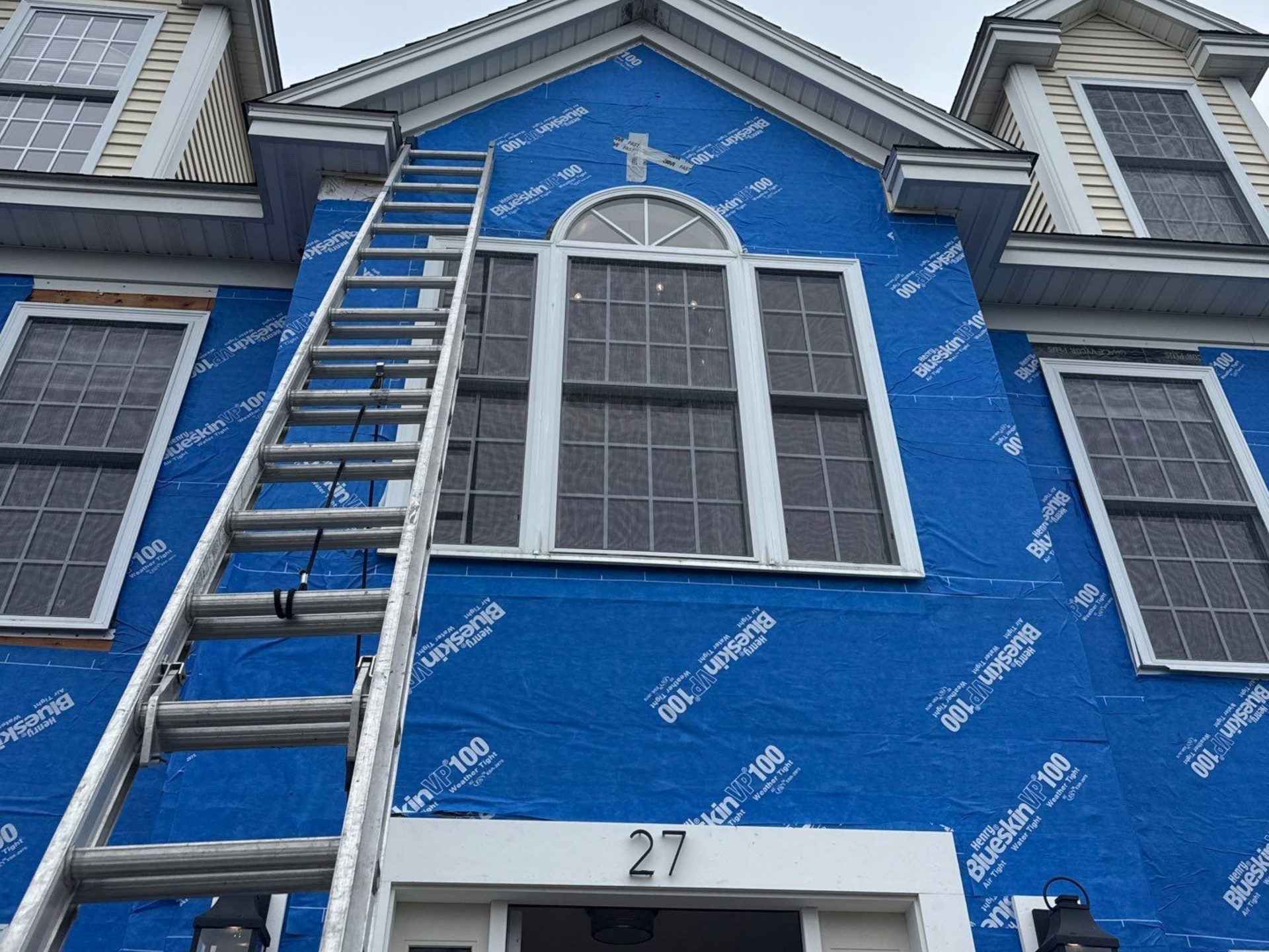 House exterior under construction, blue wrap, ladder, windows, and number 27 above the door.