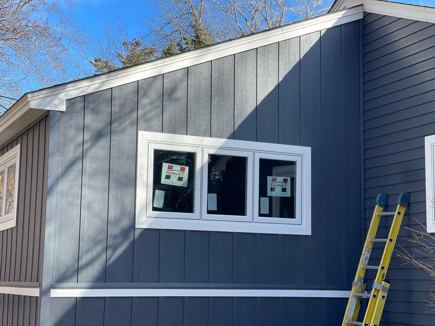 Blue siding on a house with a white-framed window. A yellow ladder leans against the wall on the right.