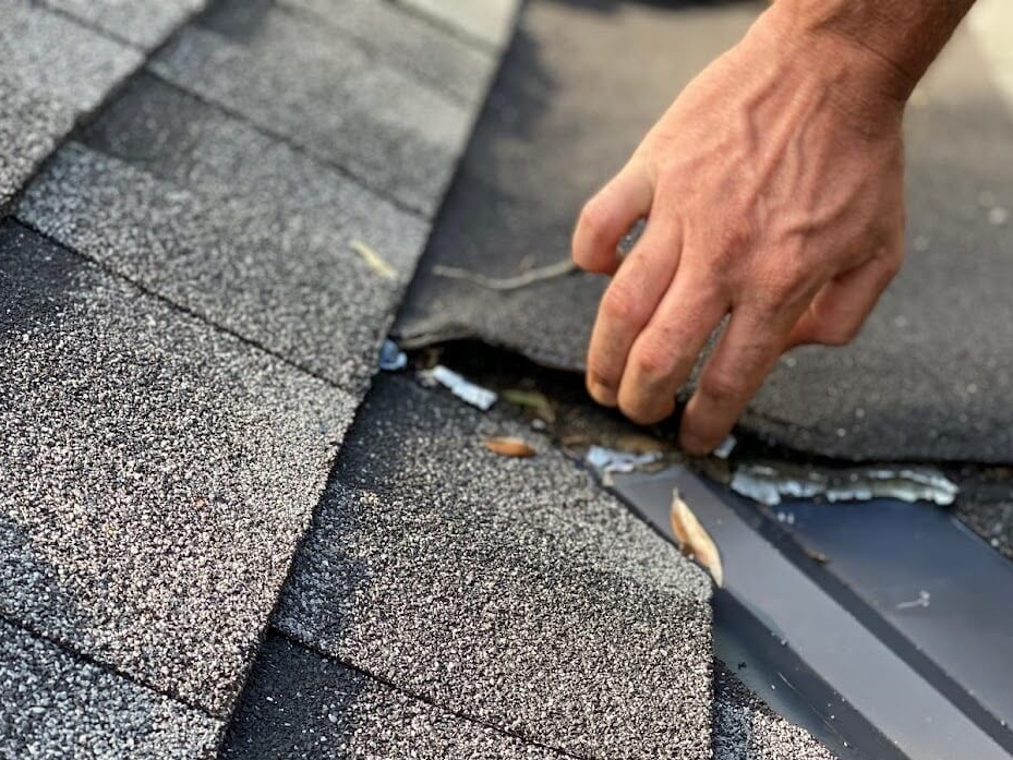 Hand examining a damaged roof near a gutter, exposing underlayment.
